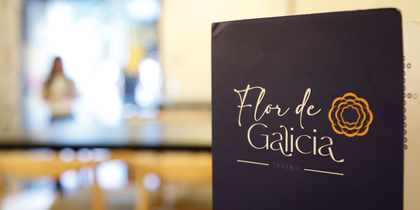 Carta de Flor de Galicia de pie sobre una mesa, con el comedor desenfocado al fondo