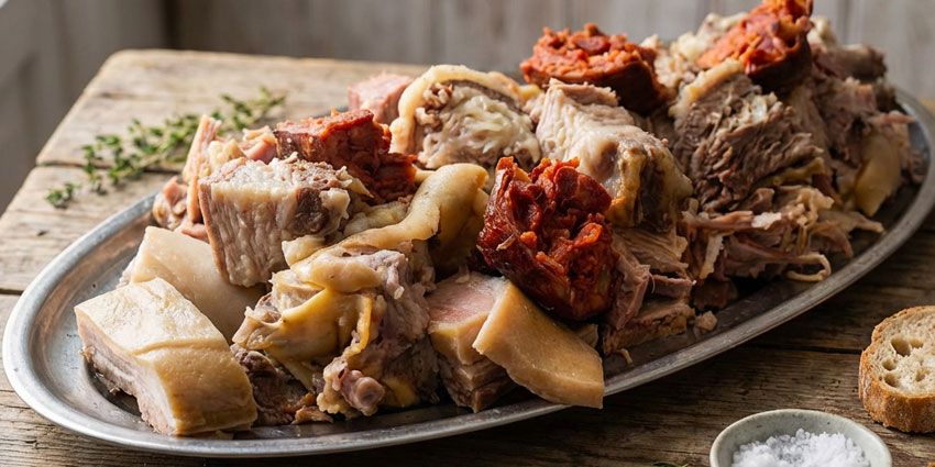 Fuente de carnes de cocido gallego con lacón, tocino y chorizo sobre una mesa de madera